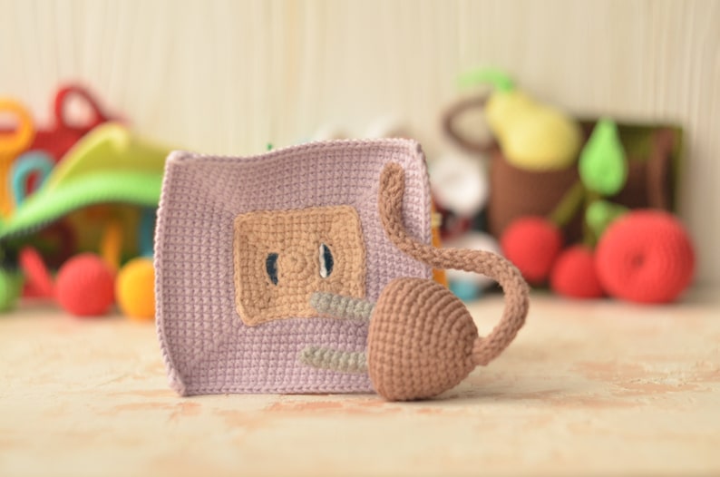 Puede incluir: Un juguete de crochet con un cuerpo morado, una cola marr&oacute;n y una cara marr&oacute;n con dos ojos. El juguete est&aacute; sentado sobre una superficie marr&oacute;n claro.