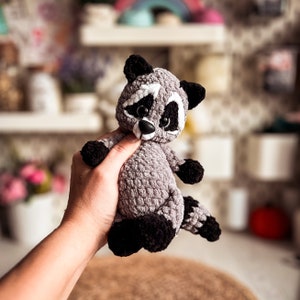 Puede incluir: Un juguete de peluche de mapache tejido a crochet con ojos negros, nariz blanca y pelaje negro y gris. El juguete est&aacute; siendo sostenido por una mano.