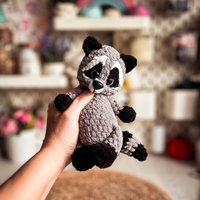 Puede incluir: Un juguete de peluche de mapache tejido a crochet con ojos negros, nariz blanca y pelaje negro y gris. El juguete est&aacute; siendo sostenido por una mano.