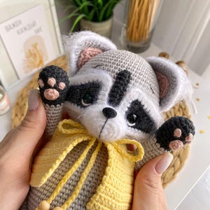 May include: A crocheted raccoon plush toy wearing a yellow crocheted vest with a bow. The toy has black eyes, a black nose, and pink ears.