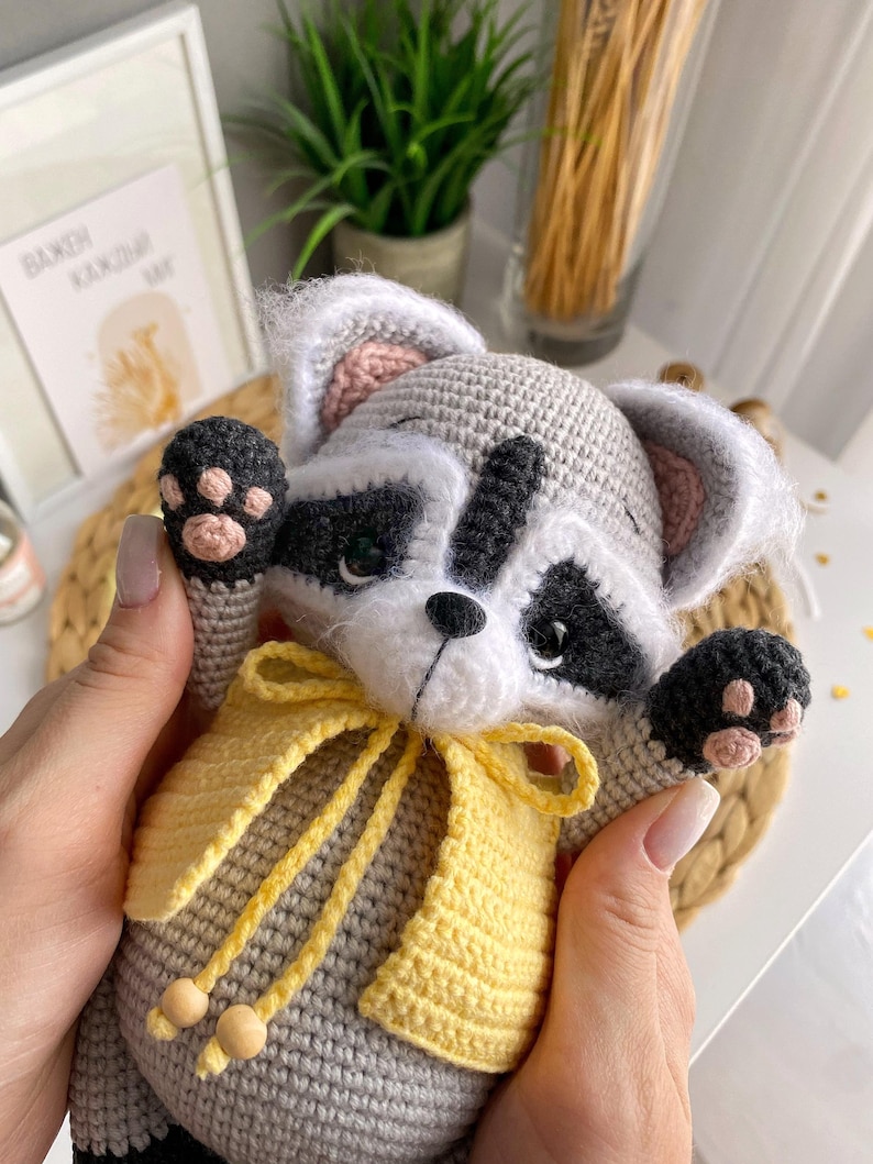May include: A crocheted raccoon plush toy wearing a yellow crocheted vest with a bow. The toy has black eyes, a black nose, and pink ears.