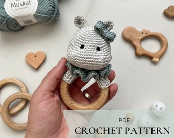 May include: Crochet pattern PDF for a gray and green hippopotamus amigurumi toy attached to a wooden teething ring. The pattern is in English.