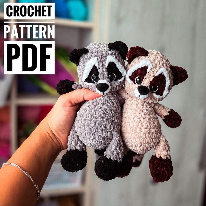 Puede incluir: Dos peluches de mapache tejidos a crochet, uno gris y otro beige, con ojos negros y detalles blancos. Los peluches se sostienen en una mano, el gris a la izquierda y el beige a la derecha.