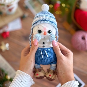 K&ouml;nnte beinhalten: Handgemachte geh&auml;kelte Schneemann-Puppe mit blau-wei&szlig; gestreifter M&uuml;tze, blauen Shorts und braunen Rentier-Stiefeln. Der Schneemann hat eine Karottennase und schwarze Augen. Die Puppe wird in den H&auml;nden einer Person gehalten.