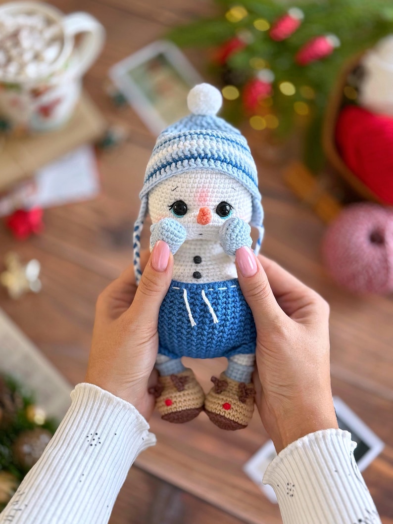 K&ouml;nnte beinhalten: Handgemachte geh&auml;kelte Schneemann-Puppe mit blau-wei&szlig; gestreifter M&uuml;tze, blauen Shorts und braunen Rentier-Stiefeln. Der Schneemann hat eine Karottennase und schwarze Augen. Die Puppe wird in den H&auml;nden einer Person gehalten.