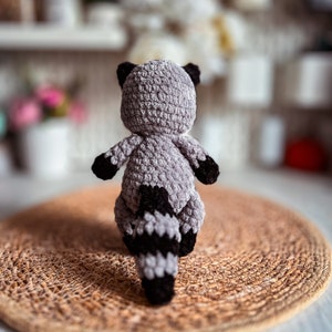 Puede incluir: Un juguete de peluche de mapache tejido a crochet con un cuerpo gris y una cola negra y gris. El juguete est&aacute; sentado en una estera de tejido marr&oacute;n.