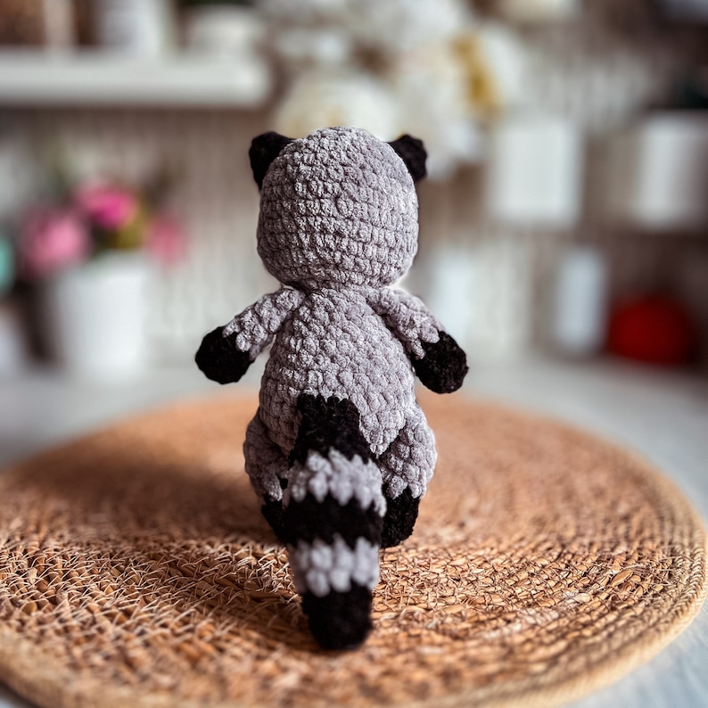 Puede incluir: Un juguete de peluche de mapache tejido a crochet con un cuerpo gris y una cola negra y gris. El juguete est&aacute; sentado en una estera de tejido marr&oacute;n.