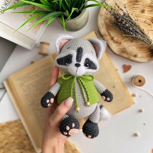 May include: A crocheted raccoon plush toy wearing a green hooded sweater. The toy has black eyes, a black nose, and a black and white striped tail.