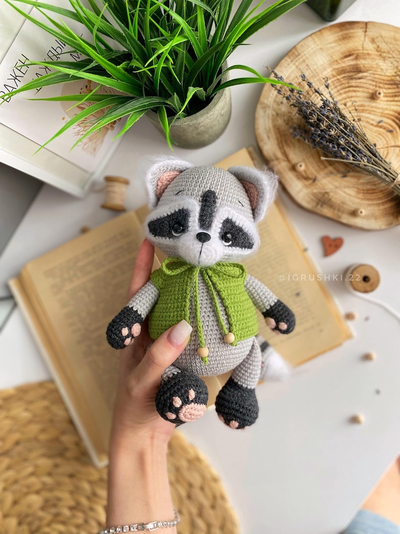 May include: A crocheted raccoon plush toy wearing a green hooded sweater. The toy has black eyes, a black nose, and a black and white striped tail.