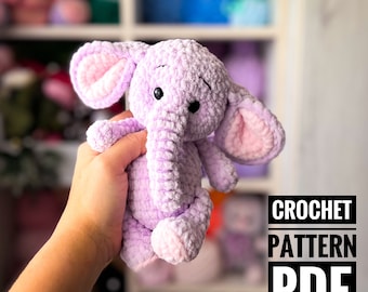 May include: A crocheted purple elephant plush toy with pink ears and a pink nose. The toy is being held in a hand. The text "CROCHET PATTERN PDF" is in the lower right corner of the image.