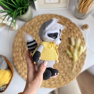 May include: A crocheted raccoon plush toy wearing a yellow shirt. The toy has black eyes, a black nose, and a black and white striped tail.