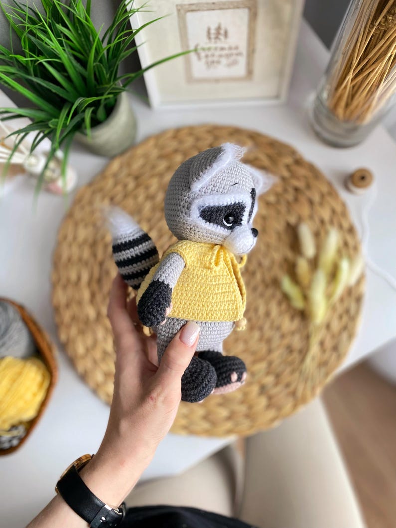 May include: A crocheted raccoon plush toy wearing a yellow shirt. The toy has black eyes, a black nose, and a black and white striped tail.