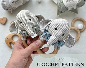 May include: Two crocheted grey elephant rattle toys with blue and green accents. The toys are attached to a wooden ring. The image includes the text "PDF CROCHET PATTERN ENG Muskat".