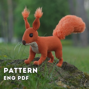 May include: A crocheted squirrel figurine with bright orange yarn, a fluffy tail, and long ear tufts. The squirrel has a cream-coloured chest, dark eyes, and thin white whiskers. The text "PATTERN ENG PDF" is visible in the lower left corner.