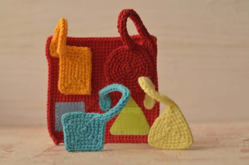 Puede incluir: Un juguete sensorial de crochet con diferentes formas y texturas de colores. El juguete est&aacute; hecho de hilo rojo, naranja, amarillo, verde, azul y gris. Tiene un lazo para colgarlo.