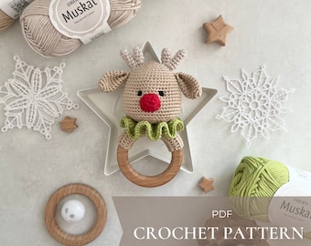 Puede incluir: Patrón de crochet PDF para un sonajero de reno. El patrón está en inglés e incluye instrucciones para hacer un reno marrón con la nariz roja y un cuello de volantes verde. El juguete está unido a un anillo de madera.