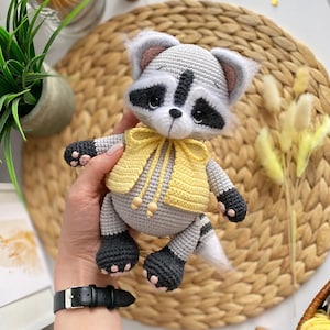May include: A crocheted raccoon plush toy wearing a yellow vest with a bow. The toy has black eyes, a black nose, and a gray and white striped tail.