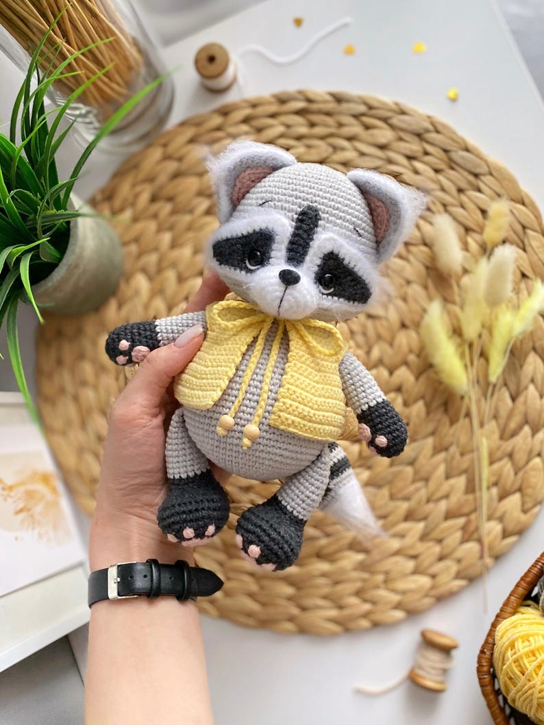 May include: A crocheted raccoon plush toy wearing a yellow vest with a bow. The toy has black eyes, a black nose, and a gray and white striped tail.