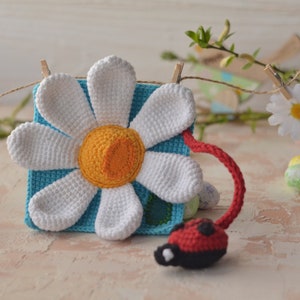 Puede incluir: Una flor de margarita de crochet con un centro azul y una mariquita unida al tallo. La margarita est&aacute; colgada de un tendedero con pinzas.