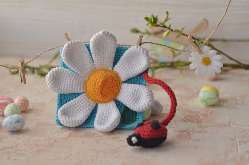 Puede incluir: Una flor de margarita de crochet con un centro azul y una mariquita unida al tallo. La margarita est&aacute; colgada de un tendedero con pinzas.
