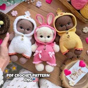 Puede incluir: Tres osos de peluche de ganchillo hechos a mano vestidos con disfraces de animales: un pato blanco, un conejito rosa y un pollito amarillo. Cada oso tiene cierres de botón y lleva un atuendo con capucha. Los osos se exhiben sobre una superficie de madera.