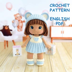 Op de afbeelding: Een handgemaakte gehaakte pop met bruin haar, blauwe ogen en een lichtblauwe jurk. De pop draagt een bijpassende hoed met berenoren en laarsjes. De tekst "CROCHET PATTERN ENGLISH PDF" is inbegrepen.