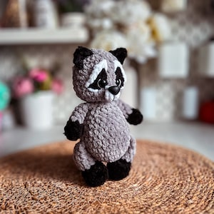 Puede incluir: Un juguete de peluche de mapache gris tejido a crochet con ojos negros y una nariz blanca. El juguete est&aacute; de pie sobre una estera tejida marr&oacute;n.