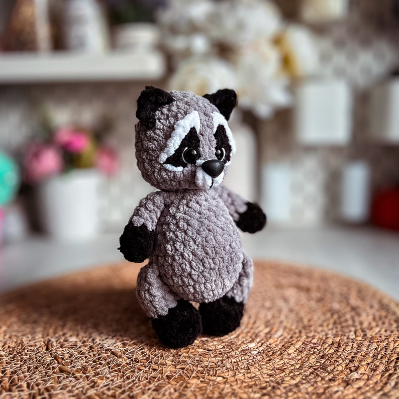 Puede incluir: Un juguete de peluche de mapache gris tejido a crochet con ojos negros y una nariz blanca. El juguete est&aacute; de pie sobre una estera tejida marr&oacute;n.