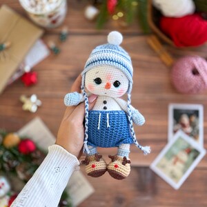 K&ouml;nnte beinhalten: Geh&auml;kelte Schneemann-Puppe mit blau-wei&szlig; gestreifter M&uuml;tze, blauen Shorts und braunen Rentierschuhen. Der Schneemann hat schwarze Augen und eine Karottennase. Die Puppe wird in der Hand gehalten. Eine festliche Weihnachtsdekoration.