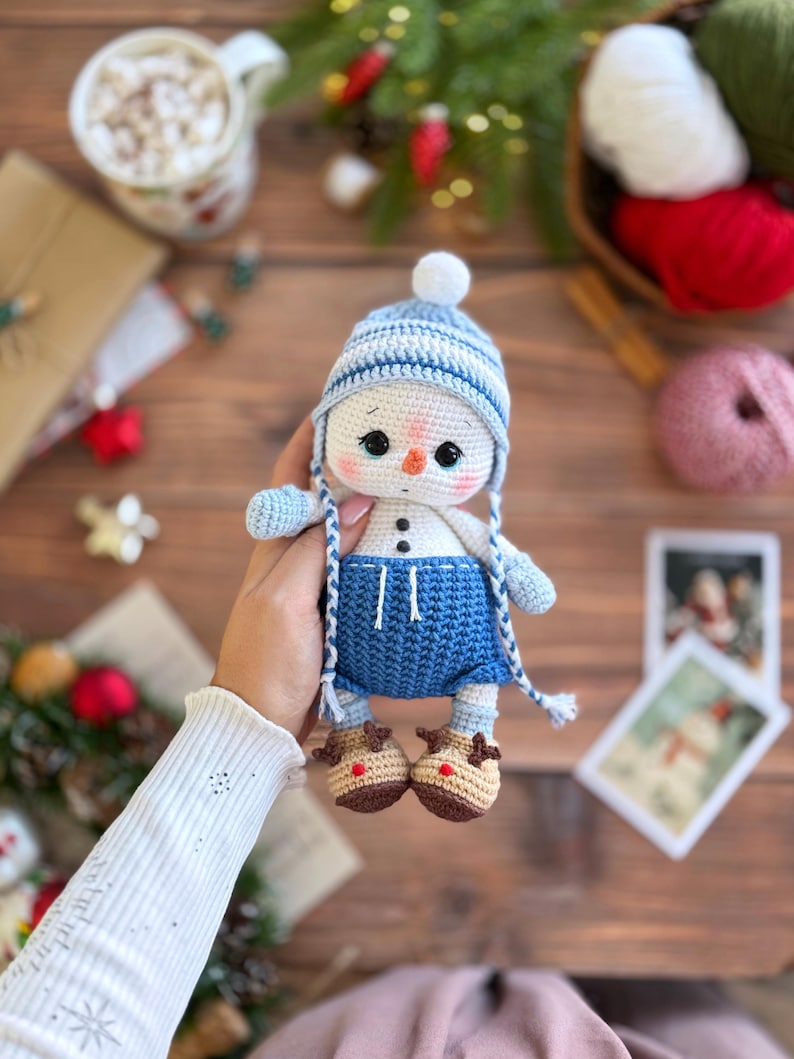 K&ouml;nnte beinhalten: Geh&auml;kelte Schneemann-Puppe mit blau-wei&szlig; gestreifter M&uuml;tze, blauen Shorts und braunen Rentierschuhen. Der Schneemann hat schwarze Augen und eine Karottennase. Die Puppe wird in der Hand gehalten. Eine festliche Weihnachtsdekoration.