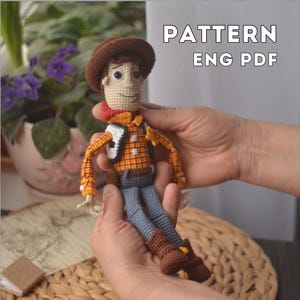 以下が含まれることがあります： 茶色の帽子、黄色のチェック柄のシャツ、青いジーンズ、茶色のブーツを履いた、手作りのカウボーイのあみぐるみ。赤いバンダナと白いベストを着ています。「PATTERN ENG PDF」の文字が画像に表示されています。