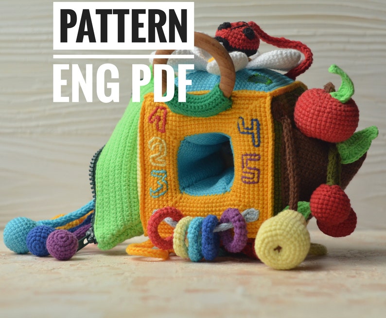 Puede incluir: Un cubo de juguete de crochet con una cremallera, un anillo de madera y una mariquita. El cubo tiene una ventana con los n&uacute;meros 1, 2, 3, 4 y 5. El juguete tambi&eacute;n tiene un sonajero, una pera y dos manzanas. El texto "PATTERN ENG PDF" est&aacute; en la parte superior de la imagen.