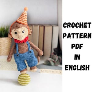 Puede incluir: Patrón de crochet PDF en inglés para un mono marrón que lleva un mono azul, un collar rojo y un gorro a rayas naranja y blanco. El mono está en equilibrio sobre una pelota verde y amarilla.
