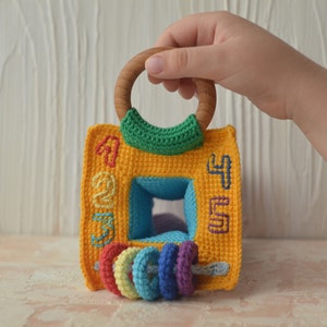 Puede incluir: Un juguete de crochet amarillo con un asa de anillo de madera y una abertura cuadrada azul. El juguete tiene los n&uacute;meros 1, 2, 3, 4 y 5 bordados en los lados. Hay cinco anillos de crochet de colores unidos a la parte inferior del juguete.