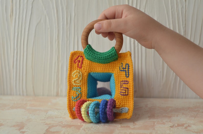 Puede incluir: Un juguete de crochet amarillo con un asa de anillo de madera y una abertura cuadrada azul. El juguete tiene los n&uacute;meros 1, 2, 3, 4 y 5 bordados en los lados. Hay cinco anillos de crochet de colores unidos a la parte inferior del juguete.