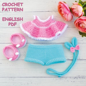 Puede incluir: Un conjunto de muñeca de ganchillo rosa y azul. El conjunto incluye un vestido rosa con ribete blanco, pantalones cortos azules, zapatos rosas y una diadema de orejas de conejo azul con un lazo rosa. El texto "CROCHET PATTERN ENGLISH PDF" es visible.