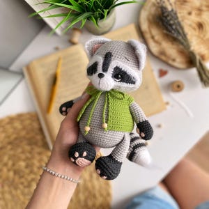 May include: A crocheted raccoon plush toy wearing a green hooded sweater. The toy has black eyes, a black nose, and a black and white striped tail.