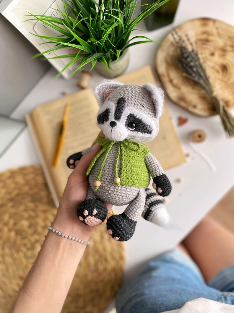 May include: A crocheted raccoon plush toy wearing a green hooded sweater. The toy has black eyes, a black nose, and a black and white striped tail.