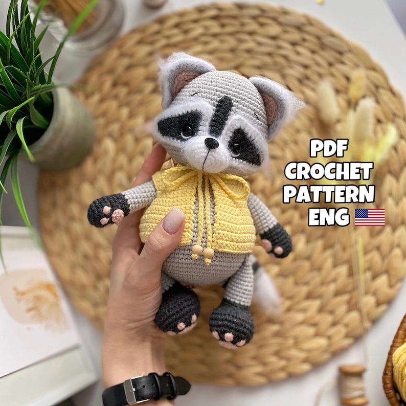 May include: A crocheted raccoon amigurumi toy wearing a yellow crocheted vest. The toy is being held by a hand. The image includes the text "PDF CROCHET PATTERN ENG".