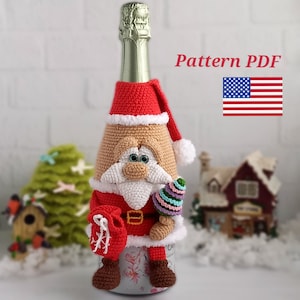 Może przedstawiać: Ręcznie robiona osłonka na butelkę w kształcie Świętego Mikołaja wykonana na szydełku w kolorze czerwonym, białym i beżowym z białą brodą i laską cukrową w ręku. Osłonka na butelkę jest zaprojektowana do standardowej butelki szampana. Wzór PDF dołączony.