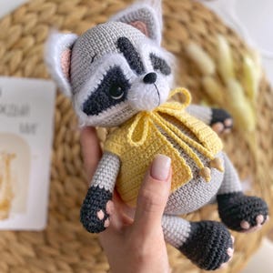 May include: A crocheted raccoon plush toy wearing a yellow crocheted vest. The toy has black eyes, a black nose, and a black and white face.