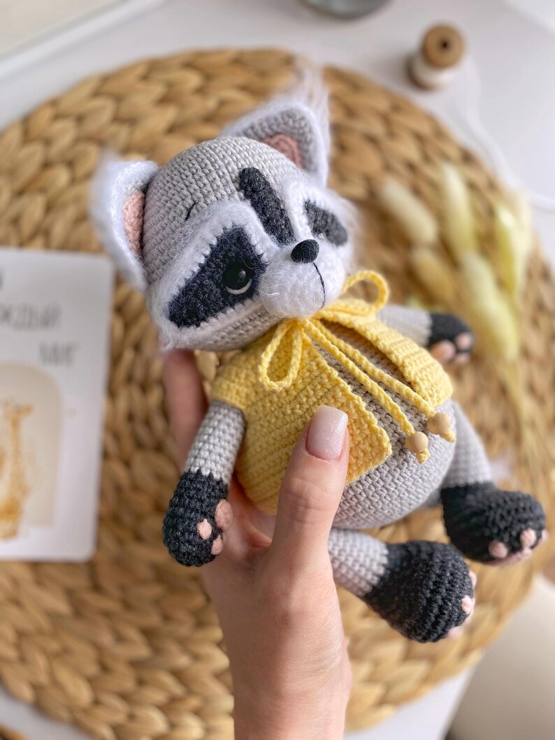 May include: A crocheted raccoon plush toy wearing a yellow crocheted vest. The toy has black eyes, a black nose, and a black and white face.