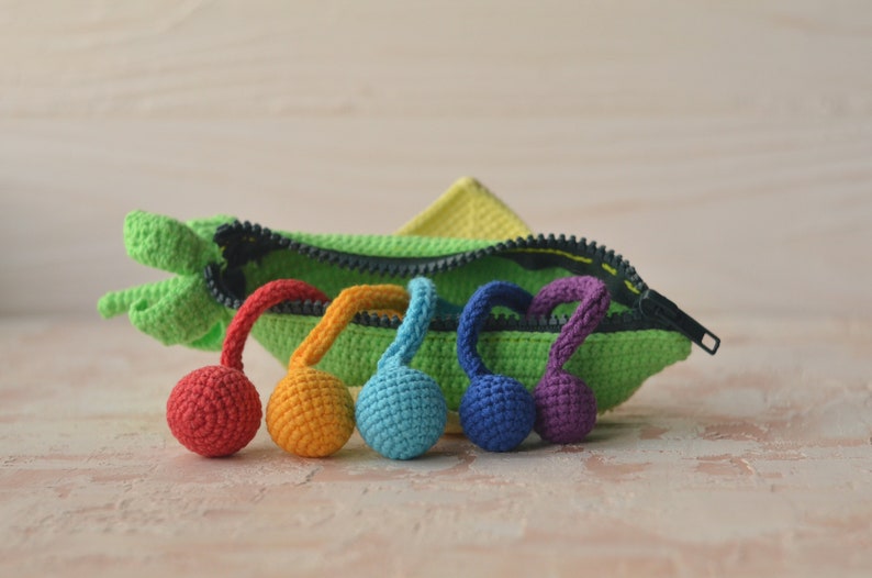 Puede incluir: Una bolsa verde de crochet con cremallera y una gama de bolas de crochet arco&iacute;ris unidas a los tiradores de la cremallera.