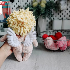 Può includere: Una bambola amigurumi di Cupido realizzata all'uncinetto con capelli in lana gialla, corpo rosa e ali bianche. La bambola tiene un piccolo ciondolo a forma di cuore argentato.