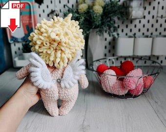 May include: A crocheted amigurumi cupid doll with a yellow yarn hair, pink body, and white wings. The doll is holding a small silver heart charm.