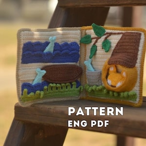 May include: A crocheted book with scenes of a lake with a creature, a boat, and a house with a fox. The book is made with various colors of yarn, including blue, green, brown, and yellow. The text "PATTERN ENG PDF" is displayed.