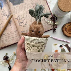 May include: A crocheted Mandragora plant in a white pot. The plant has a brown body, green leaves, and small black eyes. The pot has a label that says "Mandragora". The image also includes a book, a candle, and a crochet hook. The text "PDF CROCHET PATTERN ENG" is also visible.
