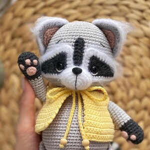 May include: A crocheted raccoon plush toy wearing a yellow vest. The toy has a gray body, black eyes, and a black nose. The toy is holding up its left paw.