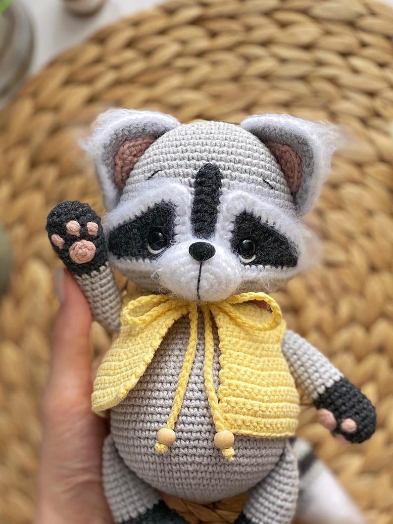 May include: A crocheted raccoon plush toy wearing a yellow vest. The toy has a gray body, black eyes, and a black nose. The toy is holding up its left paw.