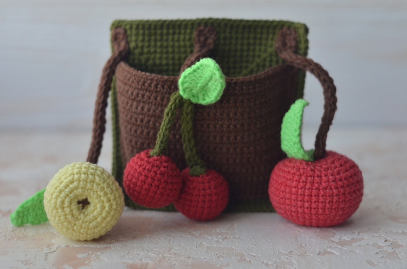 Puede incluir: Un bolsillo de pared de crochet marr&oacute;n con tres cerezas rojas de crochet y una manzana amarilla de crochet. Las cerezas y la manzana tienen hojas verdes de crochet.